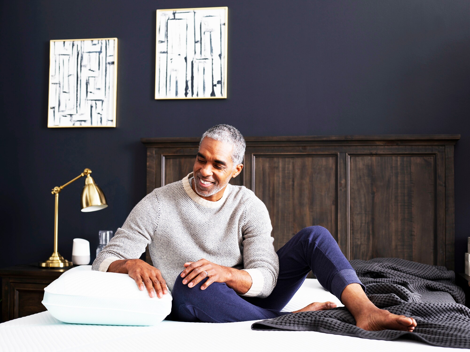 Smiling man sitting on bed with a pillow, in a dark blue room with modern decor