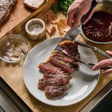 Person slicing grilled steak on a plate with sauces and seasonings nearby