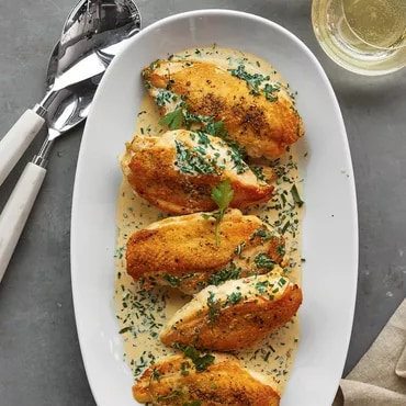 Plate of breaded chicken breasts garnished with parsley in a cream sauce, next to cutlery and a glass of wine