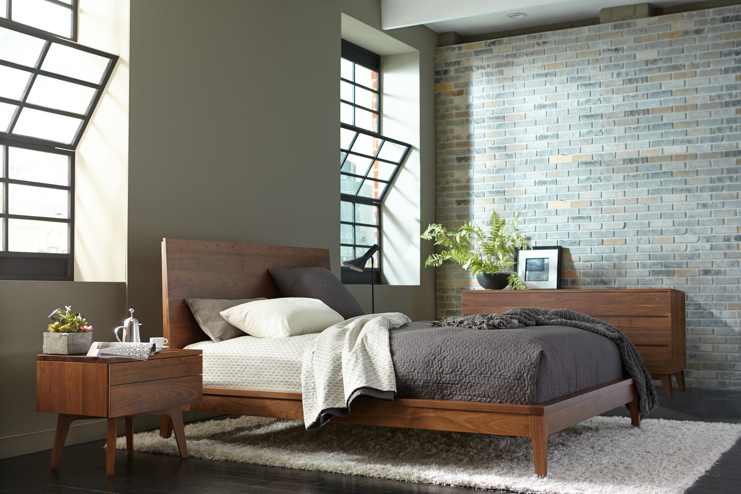Modern bedroom with a wooden bed, side tables, grey bedding, and a brick wall, illuminated by natural light