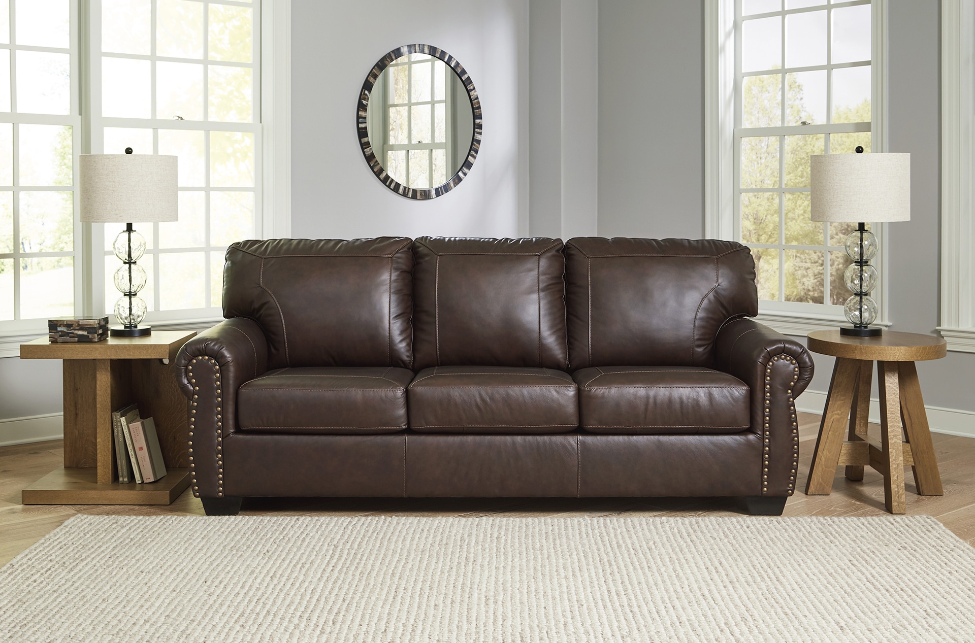 A brown leather sofa in a living room with side tables, lamps, and a round mirror