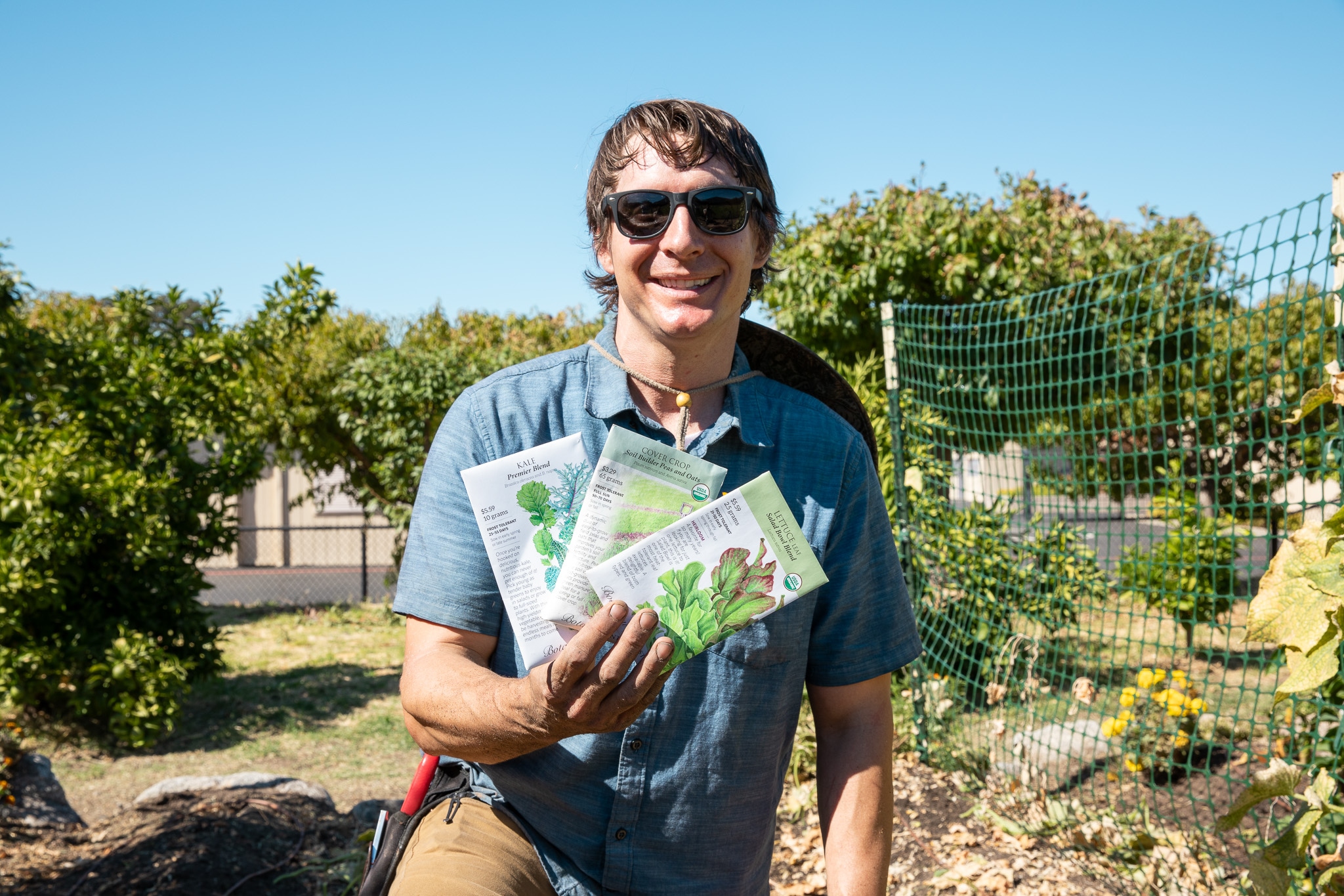 Man in sunglasses smiling and holding seed packets in a garden