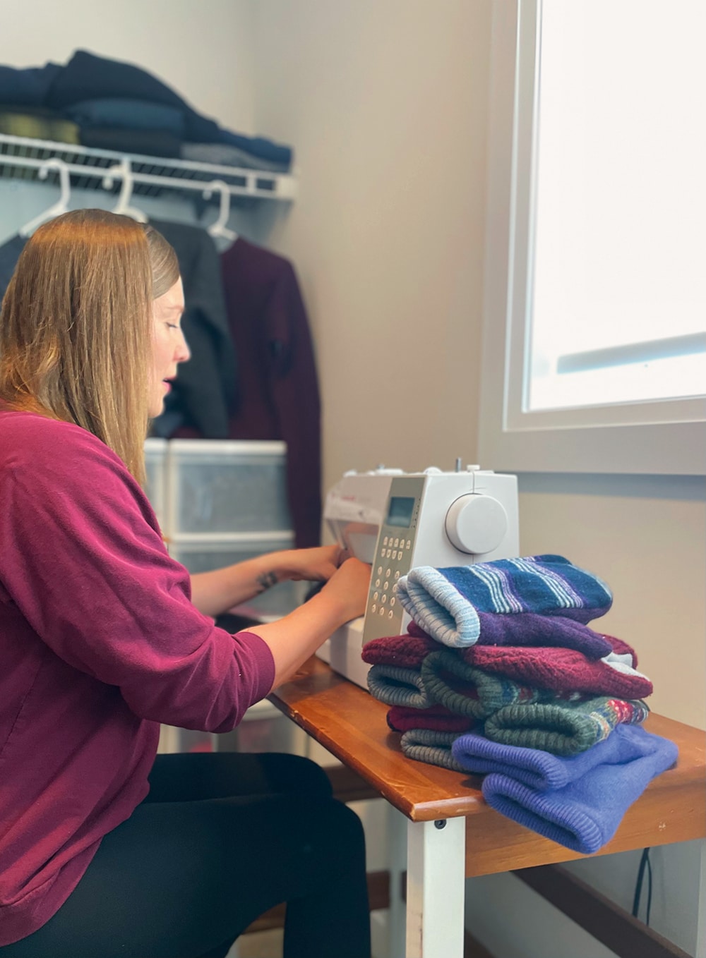 Femme utilisant une machine à coudre près d'une fenêtre, avec des piles de chaussettes colorées