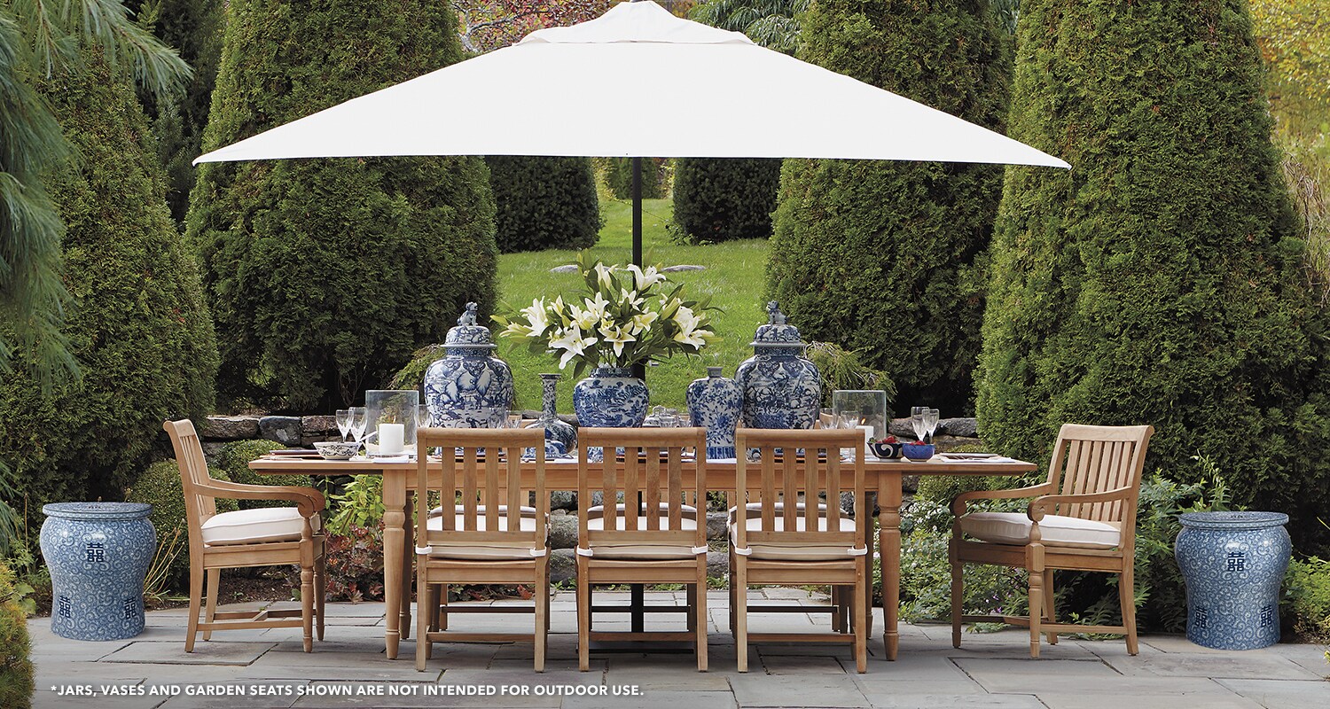 Outdoor dining area with a large table under an umbrella, surrounded by manicured hedges