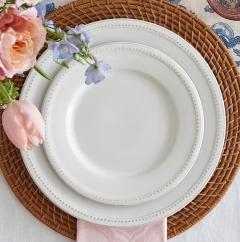 A white porcelain plate on a woven placemat with pink and purple flowers in vase