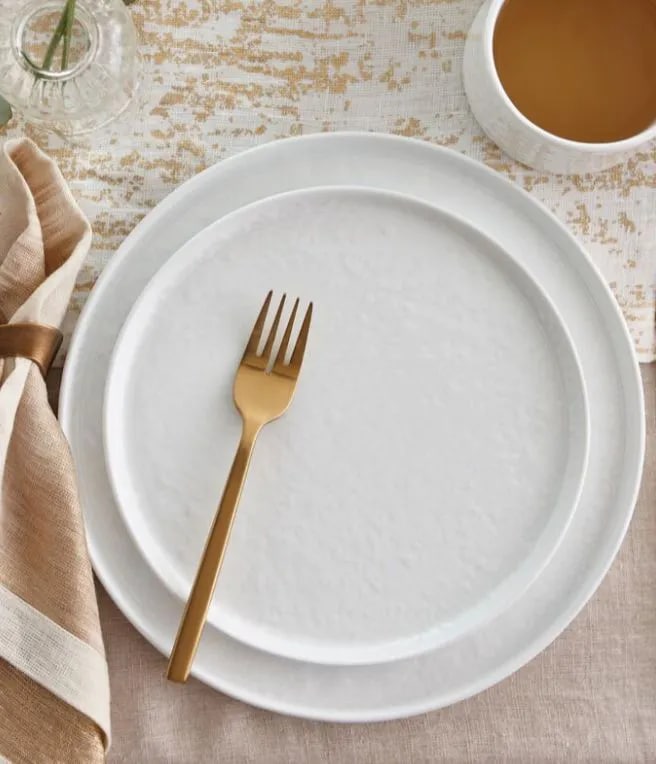 White plate with gold fork on a textured table, accompanied by a beige napkin and a bowl