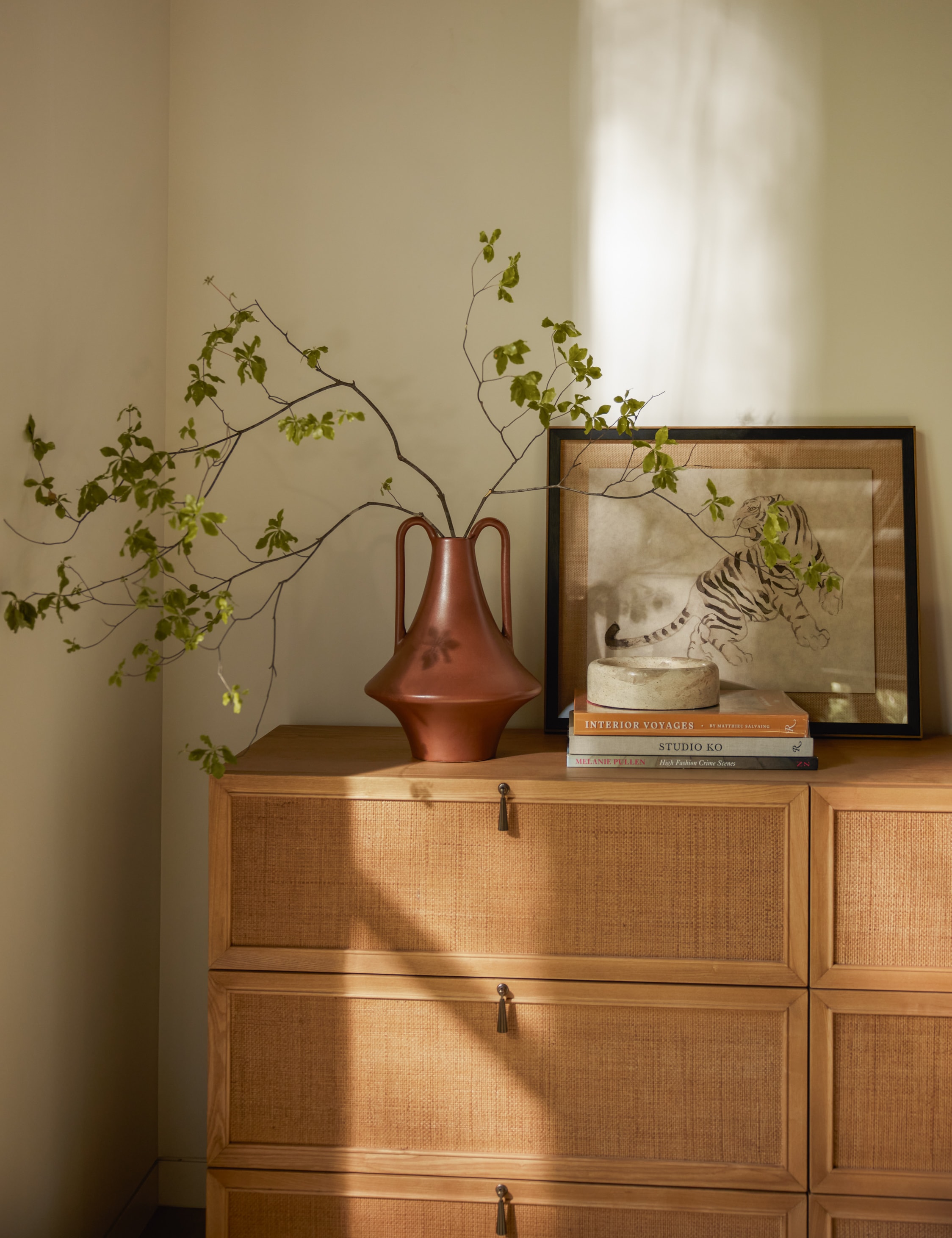 Wooden dresser with a vase of greenery, framed artwork, and books in soft sunlight