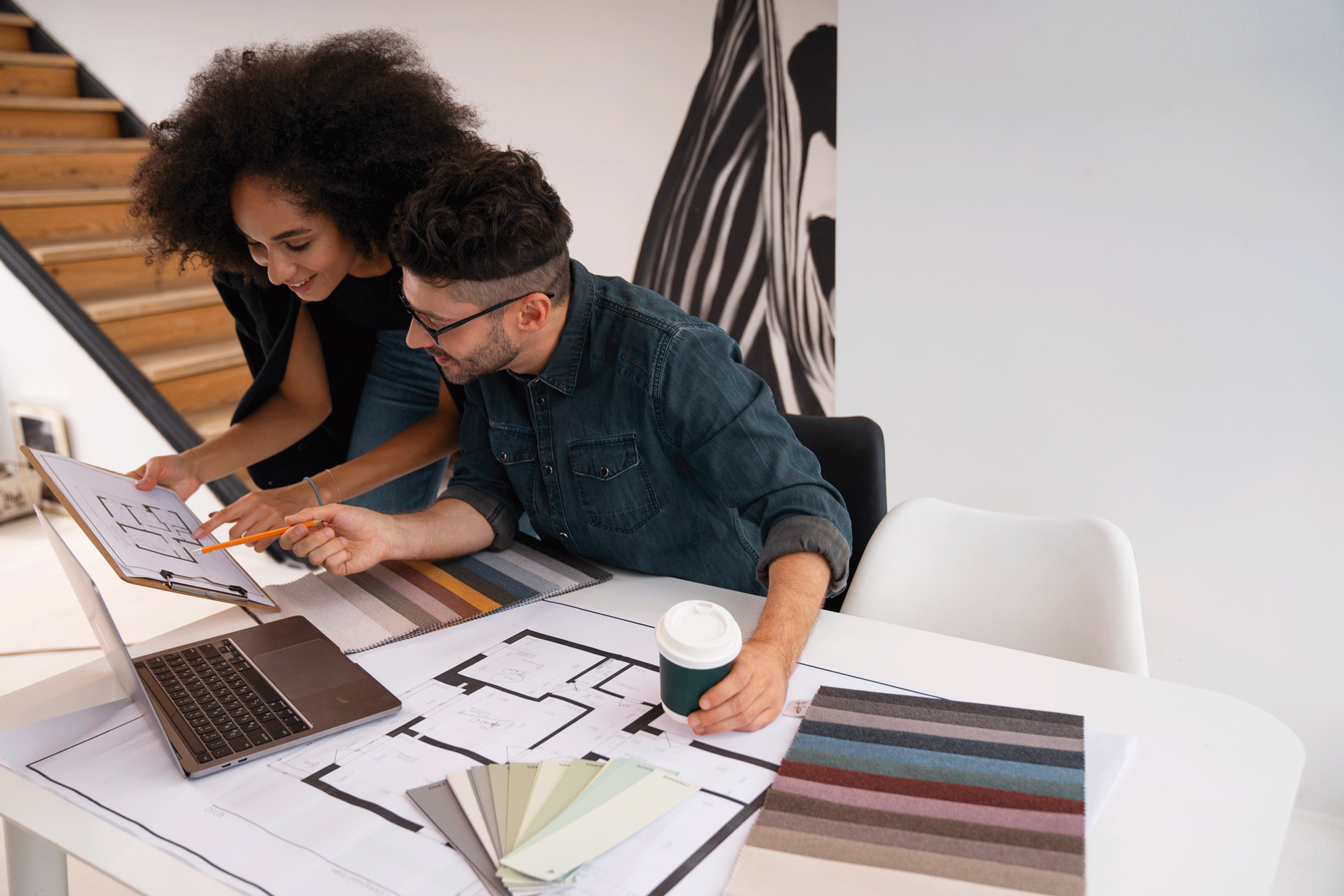 Two people reviewing documents at a desk with a laptop and color samples