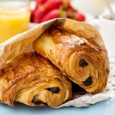 Two chocolate croissants on a plate with breakfast setting in the background