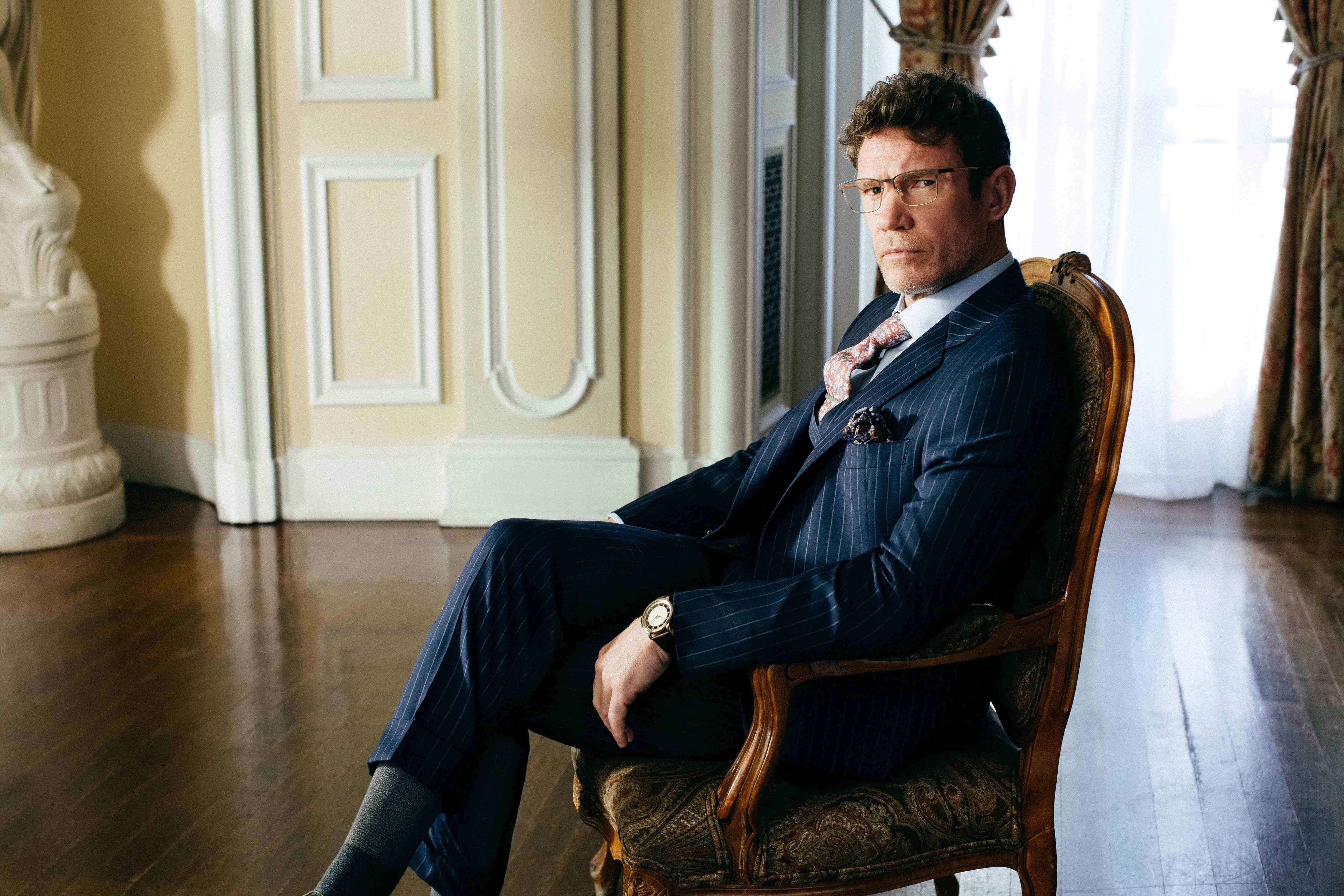 Man in a suit seated in an ornate chair in a sunlit room with classic decor