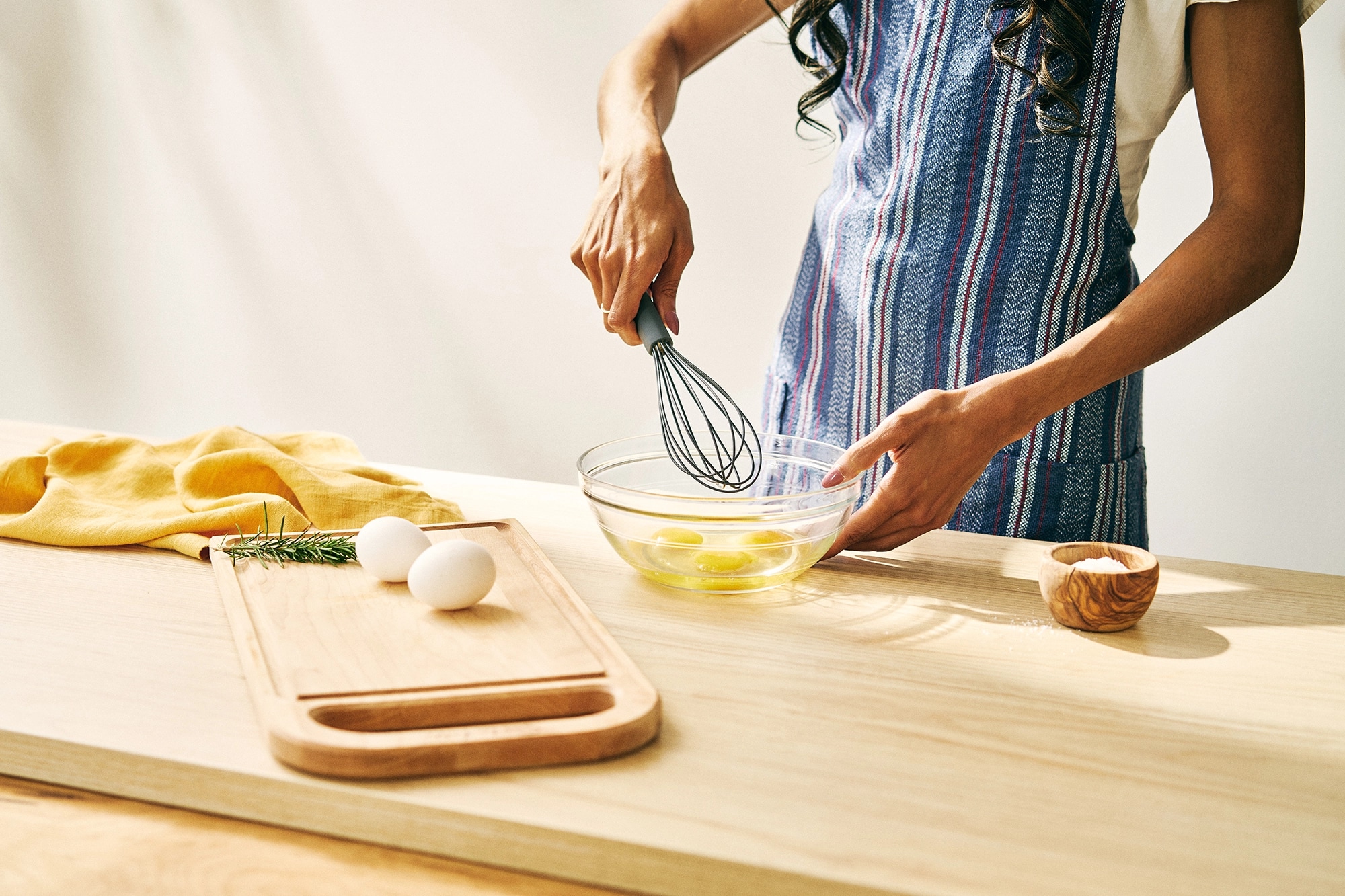 Person in a striped apron whisking eggs in a bowl on a sunny kitchen table