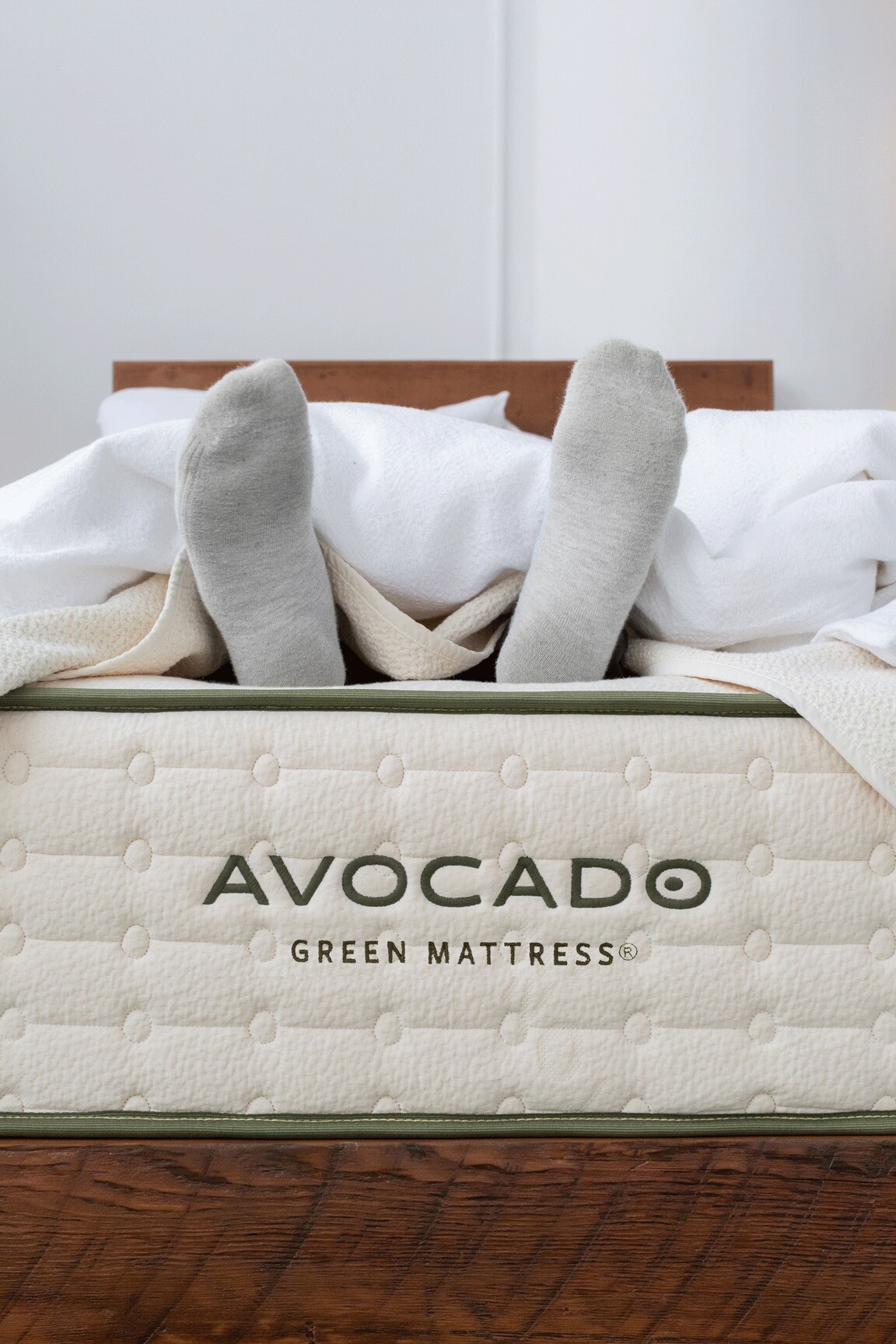 Close-up of an Avocado Green Mattress with a bed frame, white sheets, and visible feet under the covers