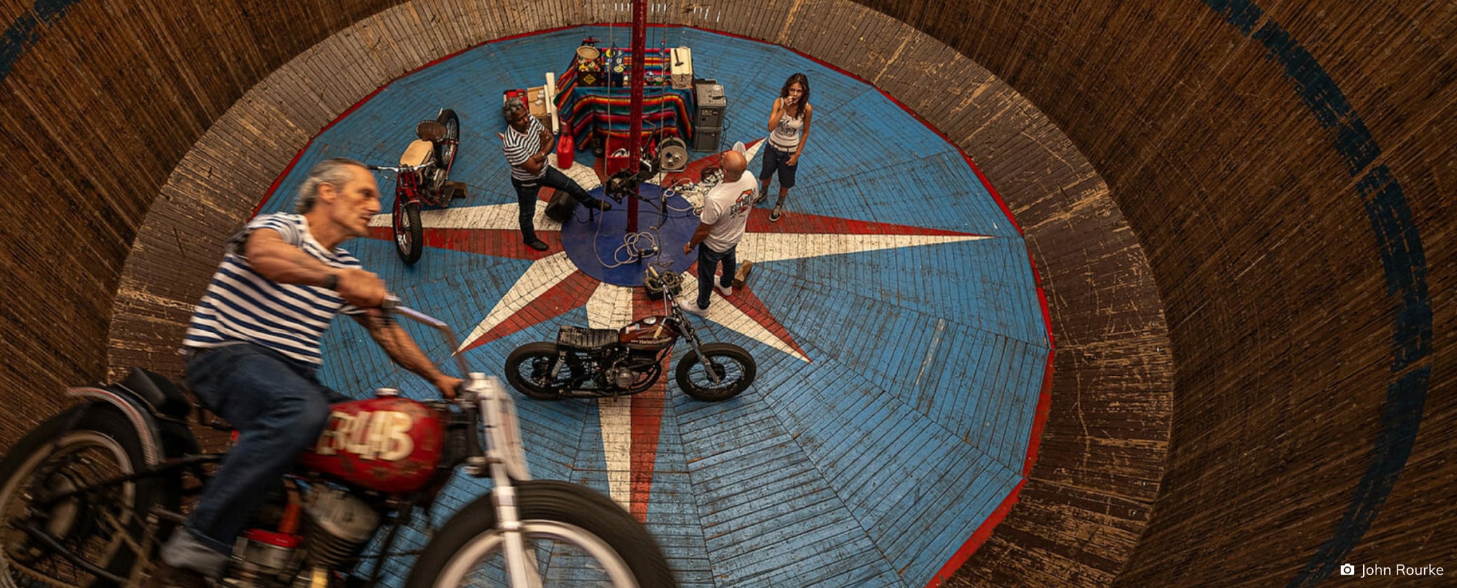 Motorcyclists performing a gravity-defying stunt inside a circular wooden wall at a carnival