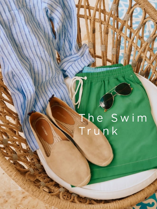 Beach essentials laid in a wicker chair: green swim trunks, blue striped shirt, loafers, and sunglasses