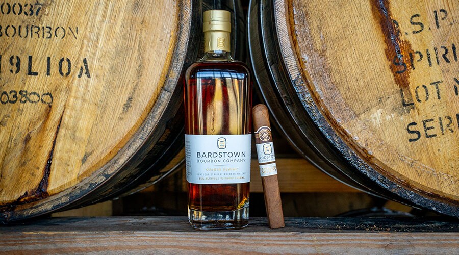 A bottle of Bardstown whisky and a cigar placed in front of aged wooden barrels